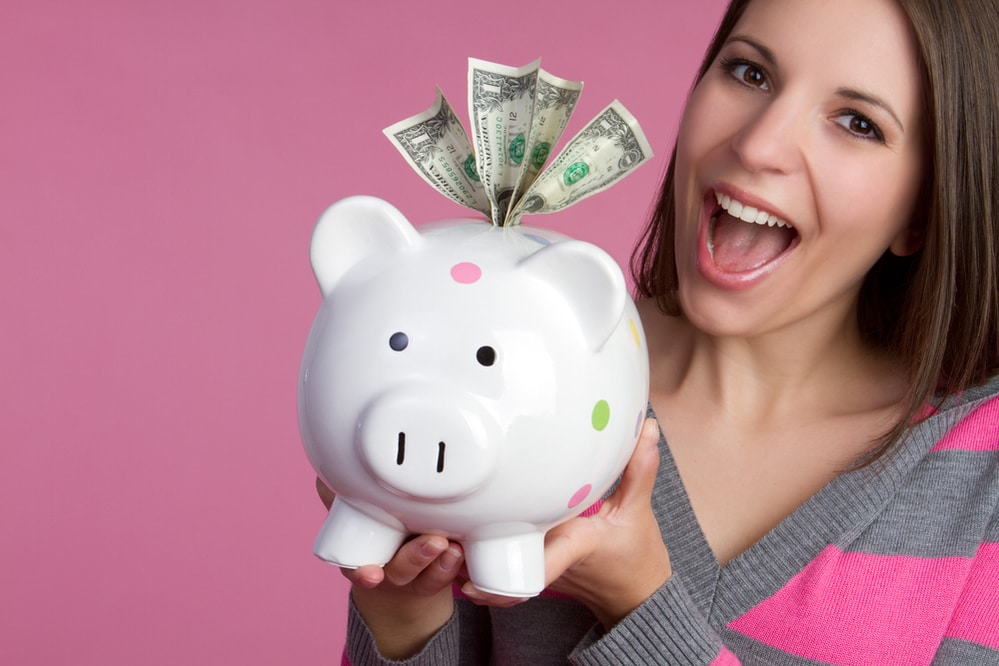 A happy young woman holding a piggy bank with money against a pink background - "How to Invest 10k" - Investment U