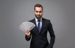 A twenty-something man in a business suit holding a fan of cash is building wealth in his twenties.